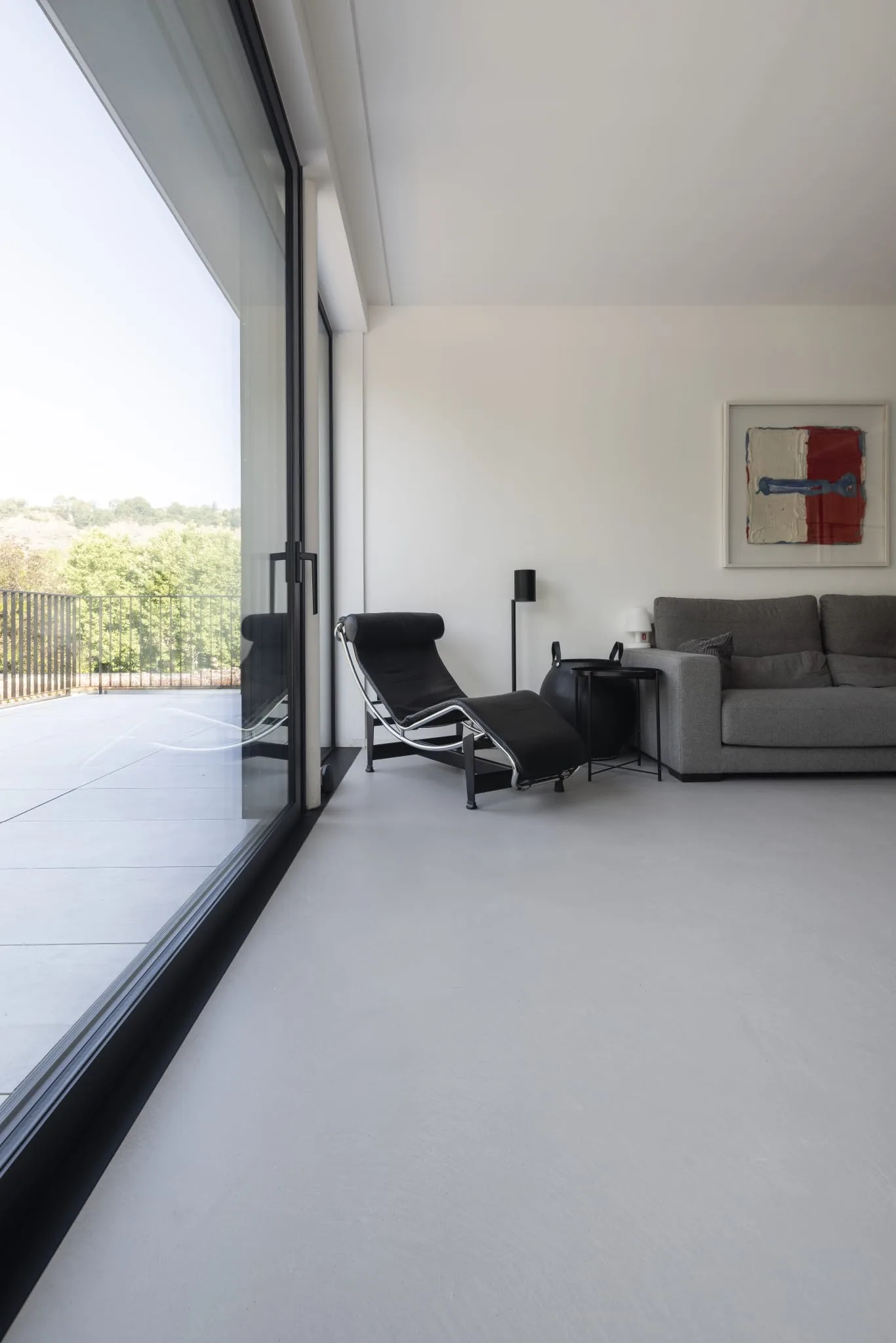 Salon épuré avec sol en béton ciré, baie vitrée, chaise longue noire et canapé gris